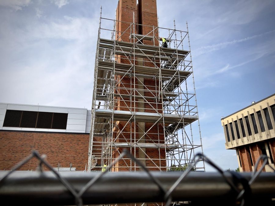 Fence view of the brick chimney undergoing construction between Buildings D and F. On top of the scaffolding near the righthand side, a construction worker stands putting down scaffolding.