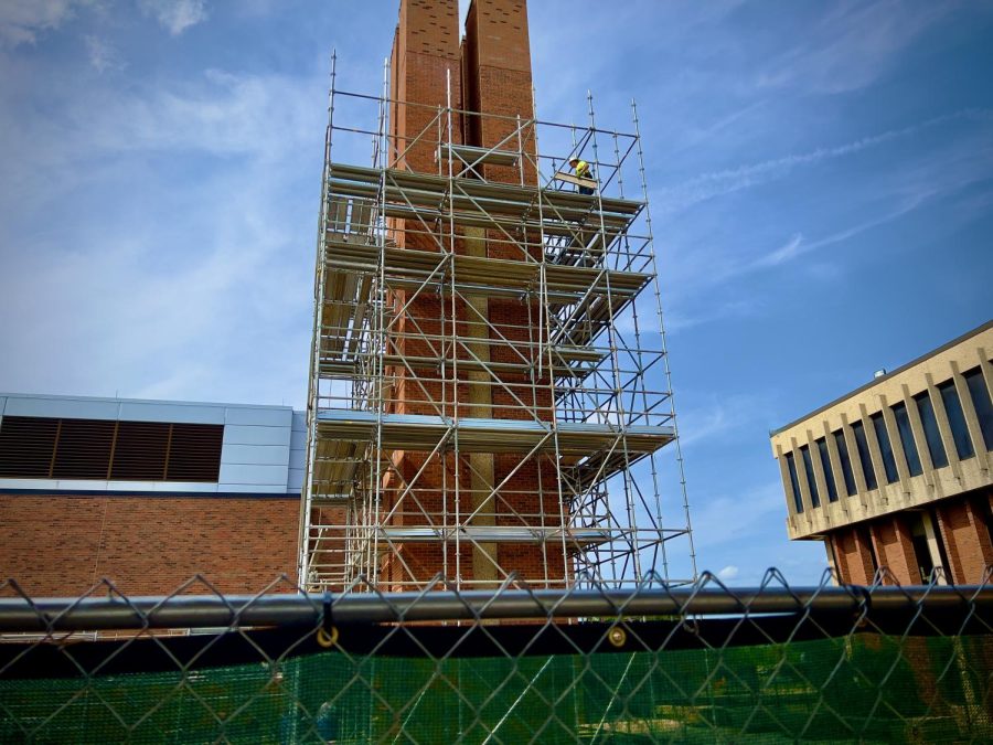 Ground view of the brick chimney undergoing construction between Buildings D and F. Top right-hand corner of the picture is a construction worker laying down pieces of scaffolding.
