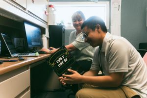 Staff-writer, Daniel Namchapassak, holds a dry erase board, saying, "First Day Of", while sitting next to Melanie Schaefer at her desk.