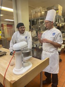 Prof. Guzzaldo in a tall white hat and apron with a Baking Operations student in a white coat and a black hat.