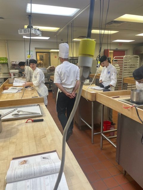 Prof. Guzzaldo walking between two rows of culinary workstations with students preparing food on wooden countertops.