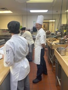 Prof. Guzzaldo smiles at a student seen from the back in a white apron and black hat.