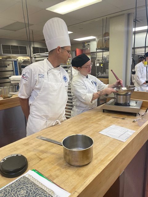 Prof. Guzzaldo in a tall white hat and apron and a student in a black cap mixing somthing in a silver bowl.