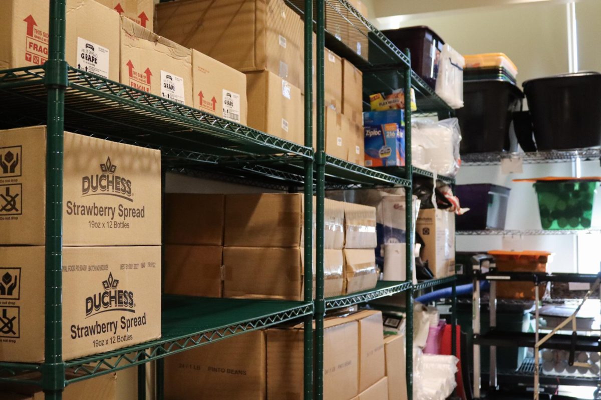 Boxes of Strawberry Spread in the stock room of the Spartan Food Pantry, on September 8, 2025. These boxes and other items are donated to the food pantry by local organizations and businesses, such as the local Sam's Club.

Alt Text: Green shelves with boxes of food and toiletries.