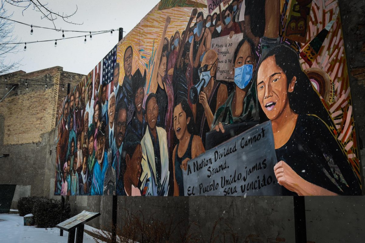 The Promise of Elgin Mural on January 30, 2025. Depicted on the right of the mural are people reacting to the events of 2020. A woman stands amongst others while holding a sign that says, "A Nation Divided Cannot Stand. El Pueblo Unido Jamas Sera Vencido". 