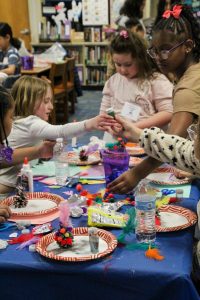 Young girls surround a table full of arts and crafts materials like feathers, pom-poms, glitter, and glue as they decorate pine-cones.