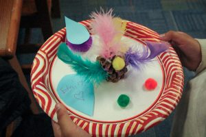 A pine cone sits on a paper plate, decorated with feathers, glitter, and pom poms. The plate is held by Emily, a young girl from Washington Elementary School.